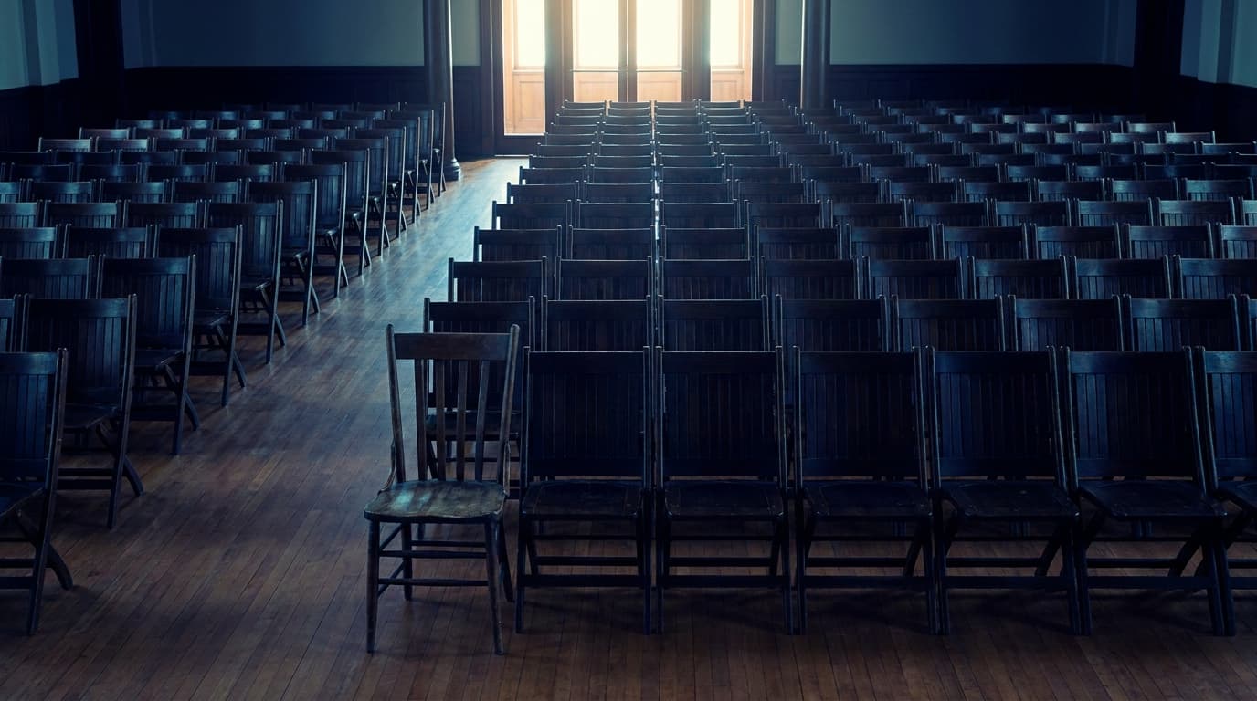 An empty aisle seat at the back of a quiet hall, with rows of chairs stretching forward towards a lit stage